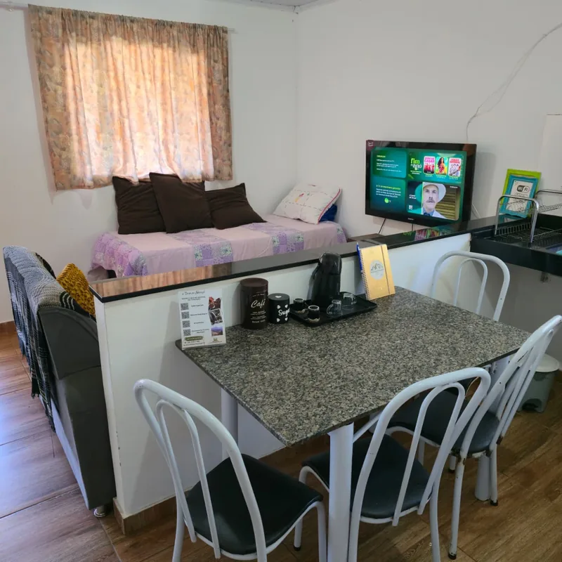 Ambiente integrado com cama, mesa de refeições, TV e cozinha ao fundo.
