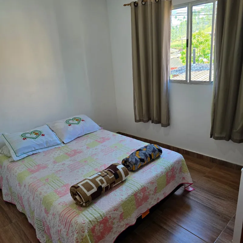 Quarto com cama de casal, colcha floral, almofadas bordadas com corações e janela com vista para o verde.