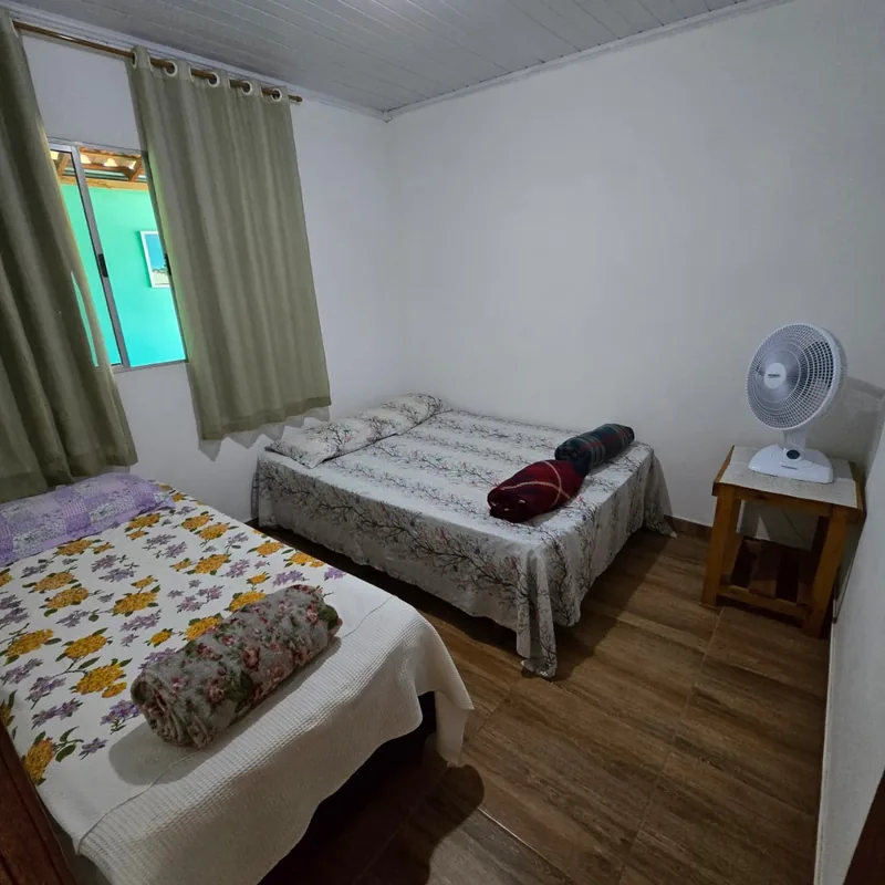 Quarto com cama de casal e cama de solteiro, criado-mudo de madeira e ventilador.