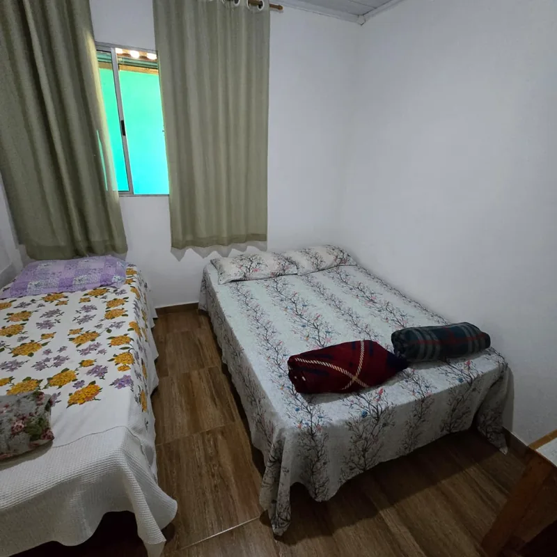 Quarto com uma cama de casal e uma cama de solteiro, colchas floridas e piso de madeira, iluminado por janela lateral.